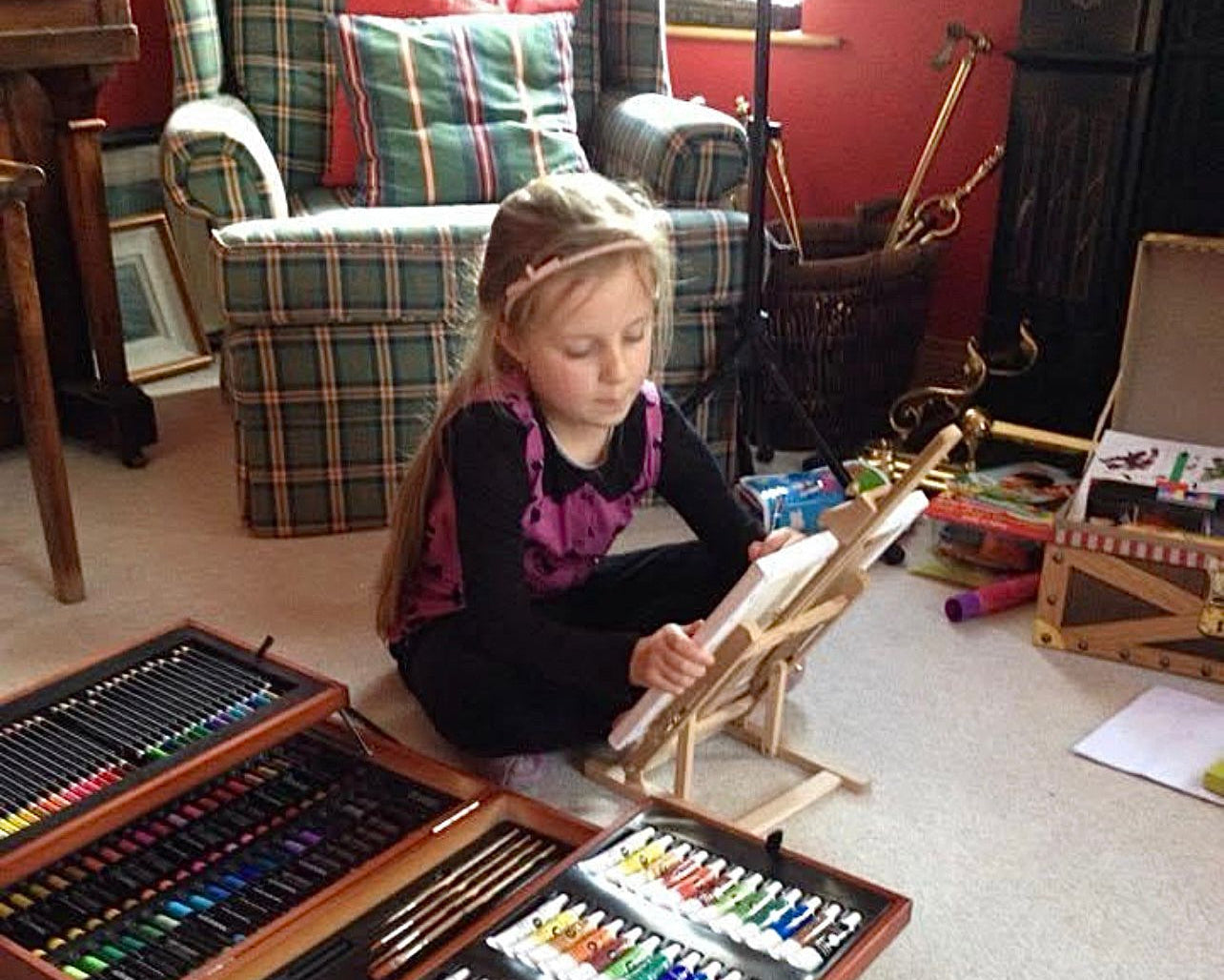 Child sitting on the floor with art supplies including a large wooden box of paints and brushes in a home setting.